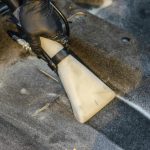 A hand in a glove using a vacuum to clean a car interior carpet, removing dirt effectively.